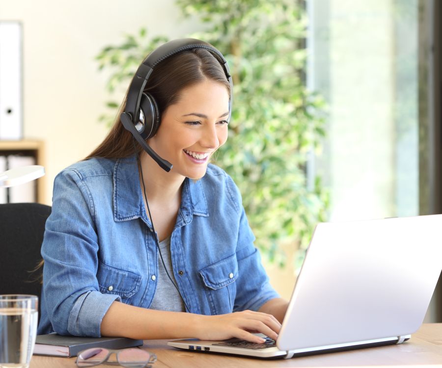 A woman wearing a headset, smiling while working on her laptop at a desk in a bright office space, with plants in the background.