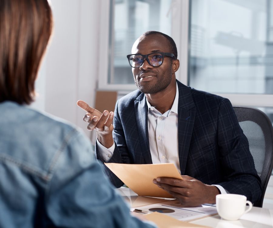 Confident smiling woman leaning against a window frame in a modern meeting room with a table full of papers and laptops