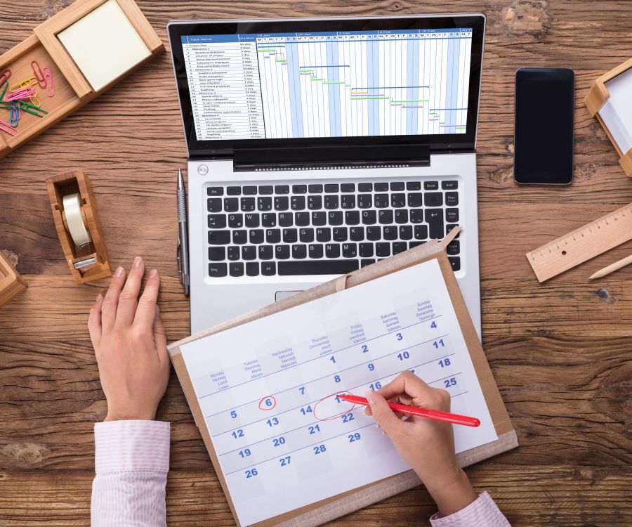 A person is writing on a calendar with a red pen. A laptop with a project management software is open nearby.